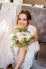 A happy bride with a bouquet in her hands sits on a soft chair in the room.