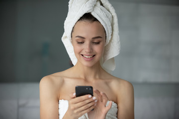 Smiling young woman in bath towel stand in bathroom using modern cellphone gadget, addicted millennial female relax after shower browsing Internet on smartphone device in bath, technology concept