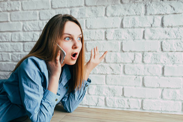 hipster teenager girl in blue shirt with long hair is talking animatedly on the phone with surprise on her face against a white brick wall and tells interesting story. young woman in loft space.