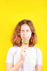 Teen girl licks a multi-colored round lollipop on yellow background