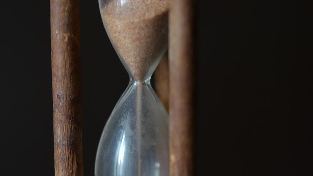 Hypnotic macro one minute running vintage hourglass on dark background. Unique wooden glass hourglass counts down the flowing sand. Countdown timer end. Time is over. Close up. B-roll for captions.
