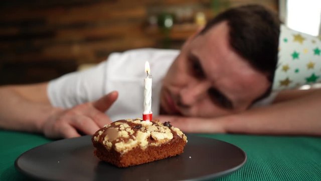 Sad lonely young man in festive hat celebrating birthday alone, sitting at the birthday cake and looking with sad eyes on it. Shooting in slow motion.