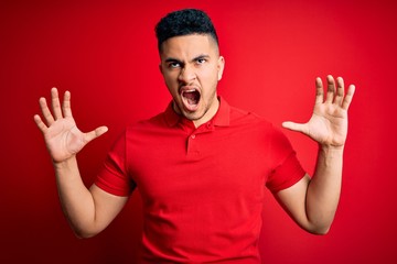 Young handsome man wearing red casual polo standing over isolated background crazy and mad shouting and yelling with aggressive expression and arms raised. Frustration concept.