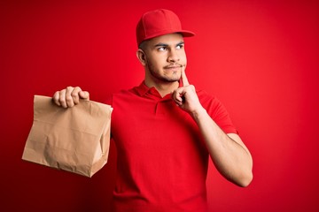 Young handsome delivery man holding paper bag with takeaway food over red background serious face...