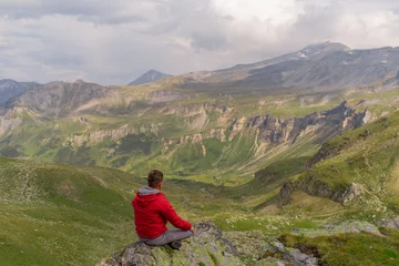 Foto op Canvas Young man in a red jacket on the stones looks at a magnificent view of the Alps mountains. © fotoplaton