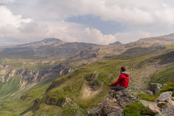 Young man in a red jacket on the stones looks at a magnificent view of the Alps mountains.