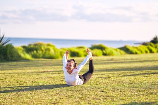 Young beautiful sportwoman smiling happy practicing yoga. Coach with smile on face teaching bow pose at park