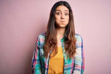 Young beautiful brunette woman wearing casual colorful shirt standing over pink background puffing cheeks with funny face. Mouth inflated with air, crazy expression.