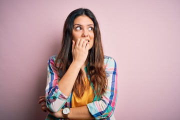 Young beautiful brunette woman wearing casual colorful shirt standing over pink background looking stressed and nervous with hands on mouth biting nails. Anxiety problem.