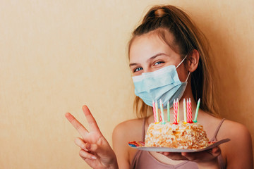 A caucasian girl in a face mask holds a birthday cake in her hands, shows a victory sign, maintains...