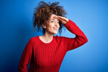 Fototapeta na wymiar Young beautiful woman with curly hair and piercing wearing casual red sweater very happy and smiling looking far away with hand over head. Searching concept.