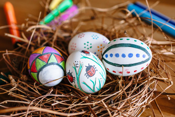Several Easter eggs, colored with a felt-tip pen, lie in a nest on a wood background. Next to it are colored markers