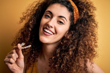 Young beautiful woman with curly hair and piercing holding dental aligner orthodontic with a happy face standing and smiling with a confident smile showing teeth