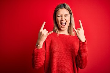 Young beautiful blonde woman wearing casual sweater over red isolated background shouting with crazy expression doing rock symbol with hands up. Music star. Heavy music concept.