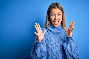 Young beautiful blonde woman wearing winter wool sweater over blue isolated background celebrating crazy and amazed for success with arms raised and open eyes screaming excited. Winner concept