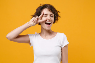 Cheerful young brunette woman girl in white t-shirt posing isolated on yellow orange background studio portrait. People emotions lifestyle concept. Mock up copy space. Blinking, showing victory sign.
