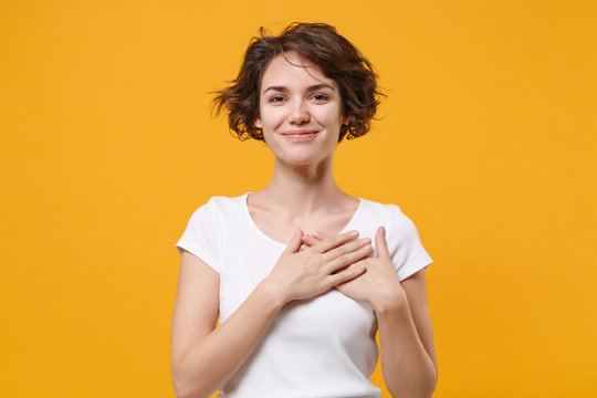 Smiling young brunette woman girl in white t-shirt posing isolated on yellow orange wall background studio portrait. People lifestyle concept. Mock up copy space. Holding hands folded on chest, heart.