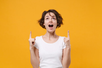Shocked young brunette woman in white t-shirt posing isolated on yellow orange wall background studio portrait. People sincere emotions lifestyle concept. Mock up copy space. Point index fingers up.