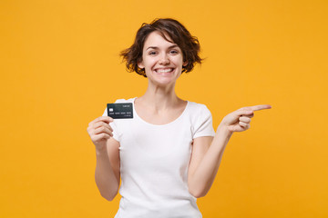 Smiling young brunette woman girl in white t-shirt posing isolated on yellow orange wall background. People lifestyle concept. Mock up copy space. Hold credit bank card, pointing index finger aside.