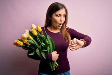 Young blonde woman holding romantic bouquet of yellow tulips flowers over pink background Looking at the watch time worried, afraid of getting late