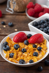 Breakfast of corn flakes with blueberries and strawberries in white bowl on wooden table.