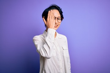 Young beautiful asian girl wearing casual shirt and glasses standing over purple background covering one eye with hand, confident smile on face and surprise emotion.