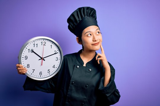 Young chinese chef woman wearing cooker uniform and hat holding clock doing countdown serious face thinking about question, very confused idea