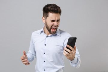 Irritated angry young unshaven business man in light shirt isolated on grey wall background. Achievement career wealth business concept. Mock up copy space. Talking on mobile phone screaming swearing.