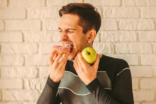 Young attractive sports man eating donut cake and holding apple in hand. Greedy man fitness coach choosing between sweet snack or healthy fruit. Fitness dieting, healthy lifestyle, weightloss