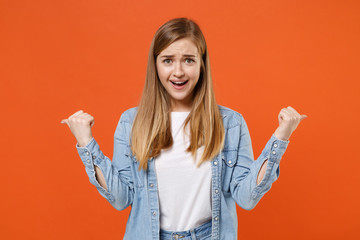 Smirked excited young woman girl in casual denim clothes posing isolated on orange background studio portrait. People sincere emotions lifestyle concept. Mock up copy space. Pointing thumbs aside.