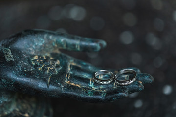 Two white golden wedding rings with diamonds on metal Buddha hand on dark background. Spiritual love concept.