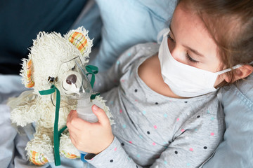Sick child recovering in bed. Little girl playing by applying medical inhalation treatment with...