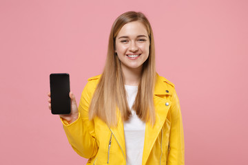 Smiling young woman girl in yellow leather jacket posing isolated on pastel pink background studio portrait. People lifestyle concept. Mock up copy space. Hold mobile phone with blank empty screen.