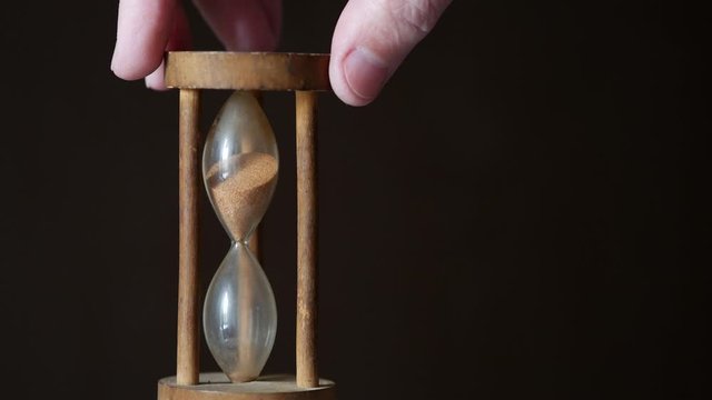 A male hand flips a vintage hourglass on a brown dark background. Unique wooden glass hourglass counts down the flowing sand. Countdown timer. Concept of passing time. B-roll for captions. Copy space