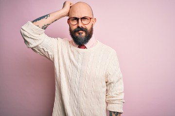 Handsome bald man with beard and tattoo wearing glasses and sweater over pink background confuse and wonder about question. Uncertain with doubt, thinking with hand on head. Pensive concept.