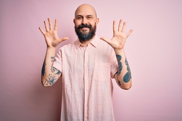 Handsome bald man with beard and tattoo wearing casual shirt over isolated pink background showing and pointing up with fingers number ten while smiling confident and happy.