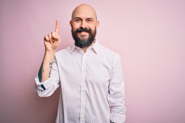 Handsome bald man with beard wearing elegant shirt over isolated pink background showing and pointing up with finger number one while smiling confident and happy.