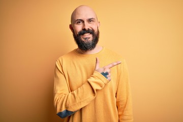 Fototapeta premium Handsome bald man with beard wearing casual sweater standing over yellow background cheerful with a smile of face pointing with hand and finger up to the side with happy and natural expression on face