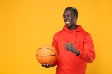 Irritated young african american man guy basketball player in red streetwear hoodie posing isolated on yellow background. Sport leisure lifestyle concept. Playing basketball hold ball clenching fist. - Powered by Adobe