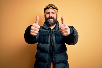 Handsome skier bald man with beard skiing wearing snow sportswear and ski goggles approving doing positive gesture with hand, thumbs up smiling and happy for success. Winner gesture.