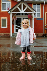Naklejka premium Cute little blonde girl in pink jacket, gray pants and rubber boots is jumping over a puddle on a rainy day