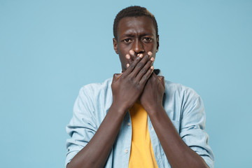 Concerned young african american man guy in casual shirt, yellow t-shirt posing isolated on blue background studio portrait. People lifestyle concept. Mock up copy space. Covering mouth with hands.