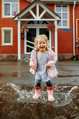 Cute little blonde girl in pink jacket, gray pants and rubber boots is jumping over a puddle on a rainy day