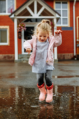 Cute little blonde girl in pink jacket, gray pants and rubber boots is jumping over a puddle on a rainy day