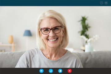 Pc screen view head shot 50s woman wear glasses looking at webcam seated on sofa at home chatting...