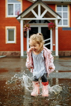 Cute little blonde girl in pink jacket, gray pants and rubber boots is jumping over a puddle on a rainy day