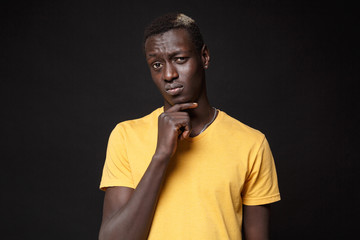 Puzzled young african american man guy in yellow t-shirt posing isolated on black background studio portrait. People sincere emotions, lifestyle concept. Mock up copy space. Put hand prop up on chin.