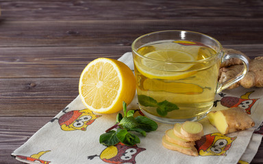 Herbal ginger tea in clear glass cup with fresh ginger, lemon and mint leaves on the linen napkin with owls, dark wooden background