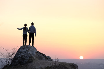 Man and woman hikers standing on big stone at sunset in mountains. Couple on high rock in evening nature. Tourism, traveling and healthy lifestyle concept.