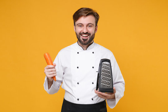 Cheerful young bearded male chef cook or baker man in white uniform shirt posing isolated on yellow background studio portrait. Cooking food concept. Mock up copy space. Holding grater and carrots.